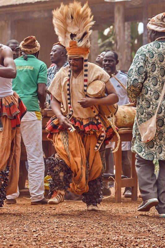 A photo of a man dressed in ethnic regalia beating w drum while dancing.